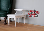 1:12 scale miniature white coffee table with lower shelf and slim legs, shown on a wood floor beside a green armchair