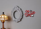 1:12 scale miniature small silver ornate oval photo frame with scrollwork, shown on a gray wall beside a table lamp