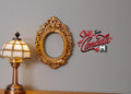 1:12 scale miniature small ornate oval photo frame in gold, shown on a gray wall beside a table lamp