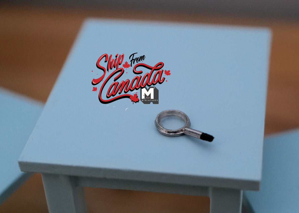 1:12 scale miniature magnifying glass with a black handle and silver rim, shown on a light blue table