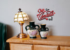 1:12 scale miniature dark green ceramic amphora vases with cream floral design, set of 2, shown on a dresser beside a lamp