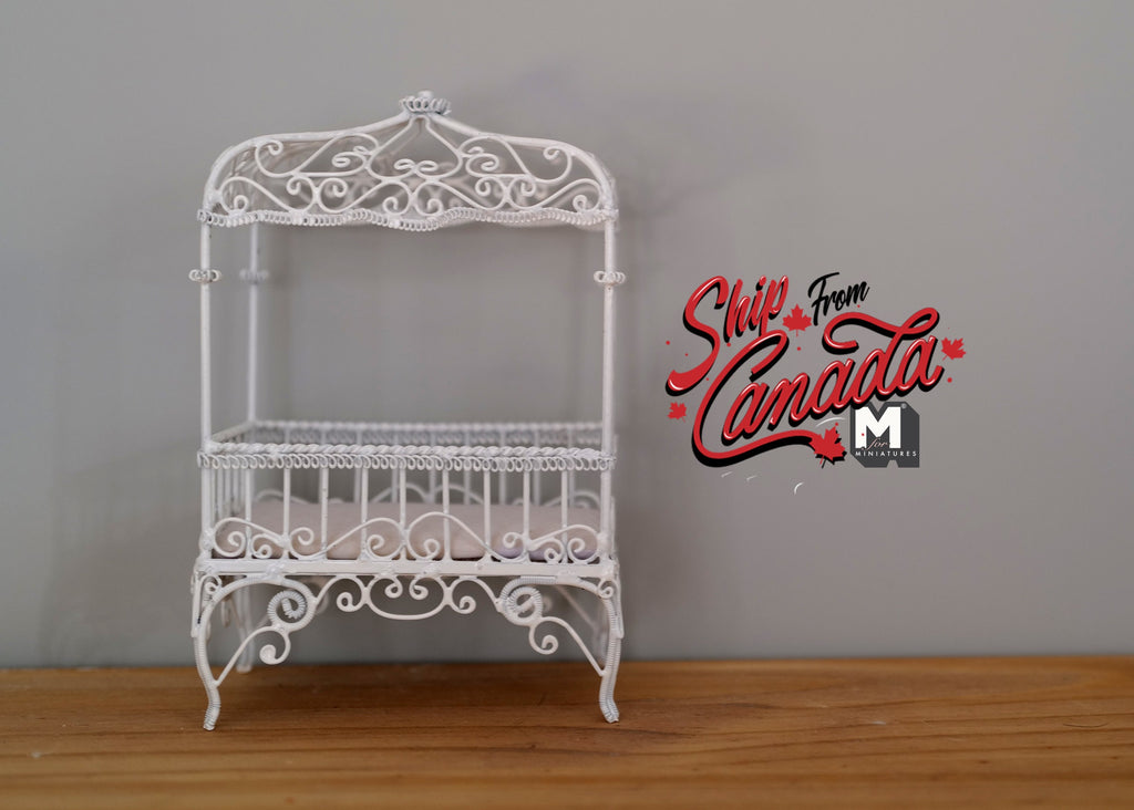 1:12 scale miniature metal baby crib in white with scroll canopy and wire sides, shown on a wood floor