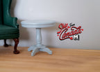 1:12 scale miniature wood round pedestal end table in light gray, shown on a wood floor beside a green armchair