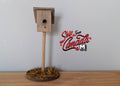 1:12 scale miniature wood birdhouse on post with round mossy base, shown on a wood surface against a gray wall