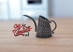 1:12 scale miniature metal watering can in dark pewter with long spout and hammered texture, on a wood table