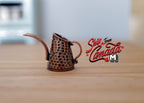 1:12 scale miniature textured metal watering can in bronze with long spout and hammered finish, on a wood surface