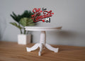 1:12 scale miniature white wood round pedestal dining table with 3 feet, shown on a wood floor beside a plant