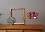 1:12 scale miniature ornate rectangular picture frame in gold with a slim open border, on a wood dresser beside a coin