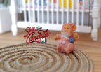 1:12 scale miniature resin baby bear figurine sitting in a pink outfit, shown on a round braided rug by a white crib