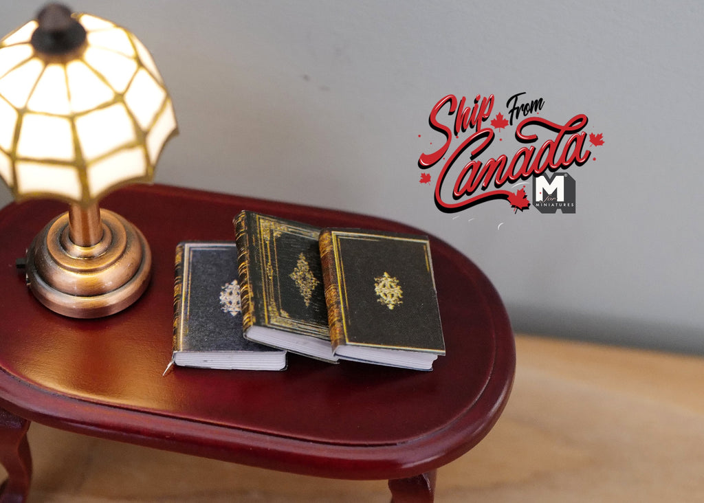 1:12 scale miniature hardcover books in dark green and black with gold cover designs, set of 3, on a table by a lamp