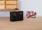 1:12 scale miniature faux leather briefcase in black with two buckle straps and top handle, on a wood table