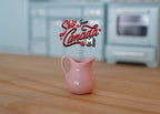 1:12 scale miniature porcelain water jug pitcher in pink with a curved handle, shown on a wood kitchen counter