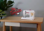 1:12 scale miniature modern sewing machine in white with red and yellow details, shown on a wood desk beside a plant