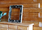 1:12 scale miniature ornate rectangular picture frame in black and gold, shown empty on a wood dresser