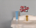 1:12 scale miniature toothpaste tube in light blue with white cap, shown standing on a white sink by a gold faucet