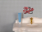 1:12 scale miniature toothpaste tube in light blue with white cap, shown standing on a white sink by a gold faucet