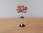 1:12 scale miniature hummingbird feeder with gold hanger, clear tube and dark base with white flower, on a wood surface