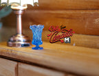 1:12 scale miniature translucent fluted vase in blue, small, shown on a wood dresser beside a lamp base