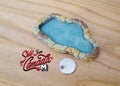 1:12 scale miniature resin garden pond with stone rim and rippled blue water, shown on a wood surface beside a coin