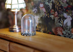 1:12 scale miniature glass dome on dark metal base, medium, shown empty on a wood dresser beside a lamp