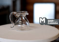 1:12 scale miniature clear glass kettle with lid, round jug body with handle and spout, shown on a round table
