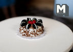 1:12 scale miniature chocolate cake with nut sides, white cream, cookie pieces and a red topper, on a round white table