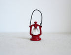 1:12 scale miniature metal hurricane oil lantern in red with black wire handle and white globe, on a light surface