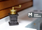 1:12 scale miniature coffee bean grinder in black and gold with crank handle and drawer, shown on a kitchen counter