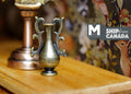 1:12 scale miniature metal amphora vase in bronze with scroll handles, shown on a wood table beside a lamp base