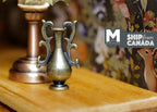 1:12 scale miniature metal amphora vase in bronze with scroll handles, shown on a wood table beside a lamp base