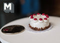 1:12 scale miniature chocolate birthday cake with white icing and red cherries on a doily, shown beside a coin
