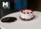 1:12 scale miniature chocolate birthday cake with white icing and red cherries on a doily, shown beside a coin