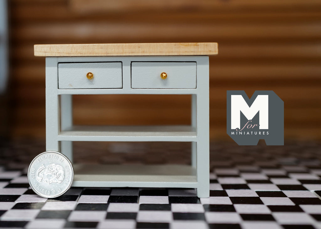 1:12 scale miniature wood kitchen island in sage green with 2 drawers, lower shelf and natural top, on a checkered floor