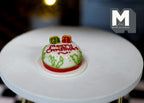 1:12 scale miniature round Christmas cake in red and white with icing, holly and gift decorations, on a white table