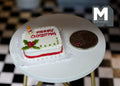 1:12 scale miniature square Christmas cake with holly, white icing with red greeting, on a white table beside a coin