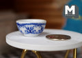 1:12 scale miniature ceramic bowl in white with blue floral pattern, shown on a white round table beside a coin