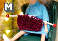 1:12 scale miniature knitting needles with a dark red knitted piece, shown on the lap of a doll in a green armchair