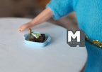 1:12 scale miniature chocolate brownies with a green garnish in a light blue dish, shown on a white table by a doll hand