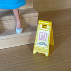 1:12 scale miniature folding caution floor sign in yellow, A frame style, shown on a wood floor beside stairs