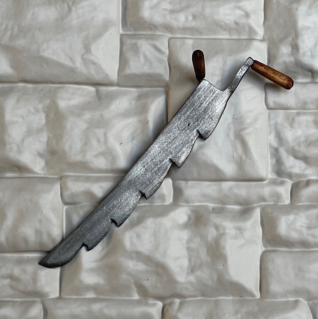 1:12 scale miniature metal hay knife with serrated blade and wood handles, shown leaning on a white stone wall