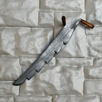 1:12 scale miniature metal hay knife with serrated blade and wood handles, shown leaning on a white stone wall