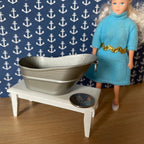 1:12 scale miniature metal baby bathtub in gray pewter tone, shown on a white bench beside a doll and a coin