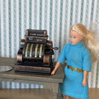 1:12 scale miniature metal antique style cash register, large, copper tone with ornate keys, shown beside a doll