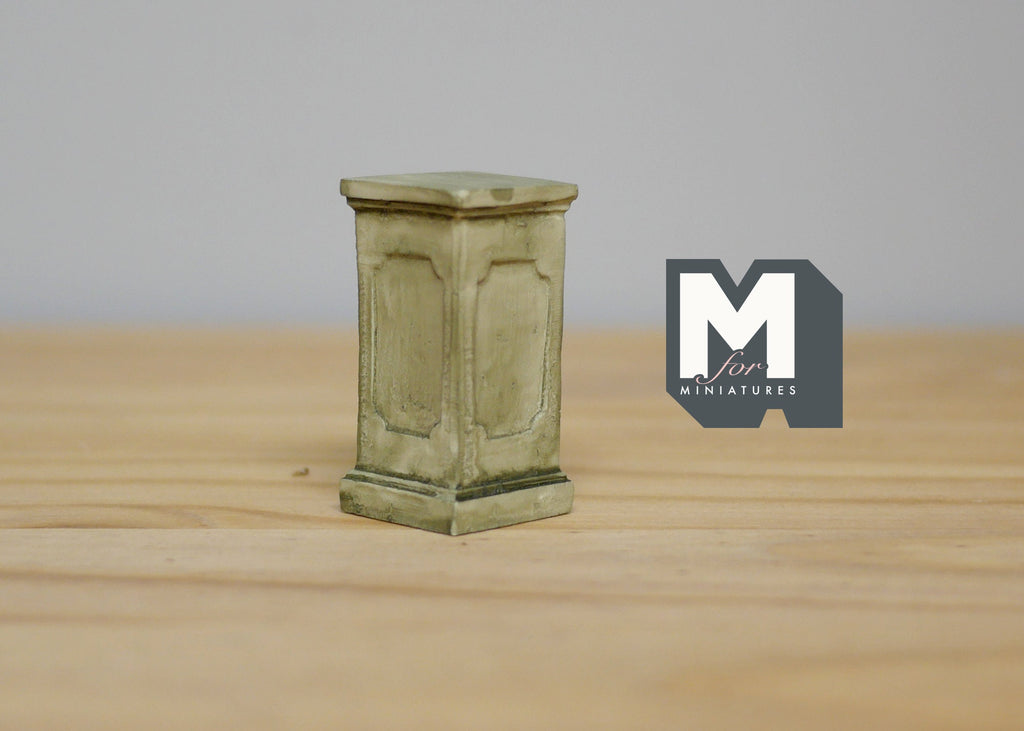 1:12 scale miniature square garden pedestal for plants in gray with paneled sides and a weathered finish, on a wood table