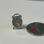 1:12 scale miniature metal twin bell alarm clock in silver with Roman numeral face, shown beside a coin for scale