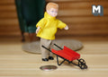 1:12 scale miniature metal child's wheelbarrow in red and black, shown with a figure in a yellow coat and a coin for scale