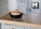 1:12 scale miniature metal cooking pot in black with beige lid and side handles, shown on a wood kitchen counter