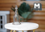 1:12 scale miniature clear glass decanter with stopper, tall bottle shape, shown on a round white marble look table