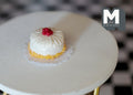 1:12 scale miniature cherry cake with white frosting, red cherry on top and yellow crumb edge, on a white table
