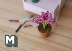 1:12 scale miniature potted pink and purple striped leaf plant in a clay pot, on a wood floor beside garden tools